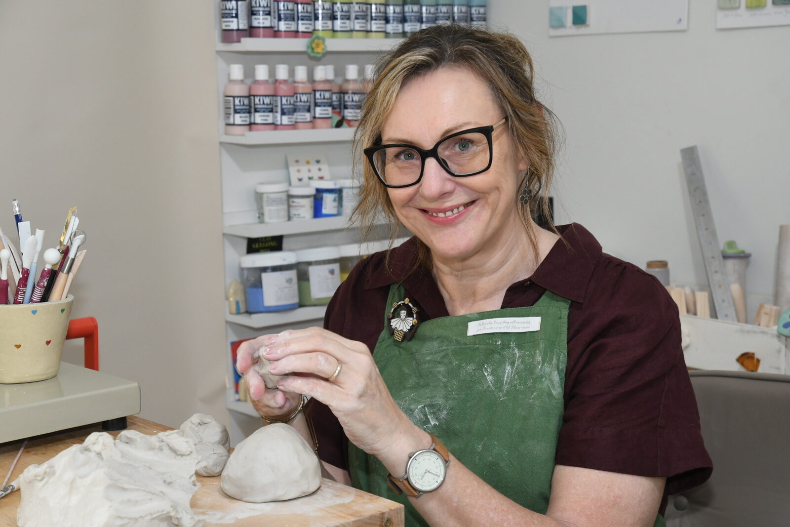 Dani Green in her studio, shaping a piece of clay at the workbench with modelling tools and shelves of glaze bottles behind her.