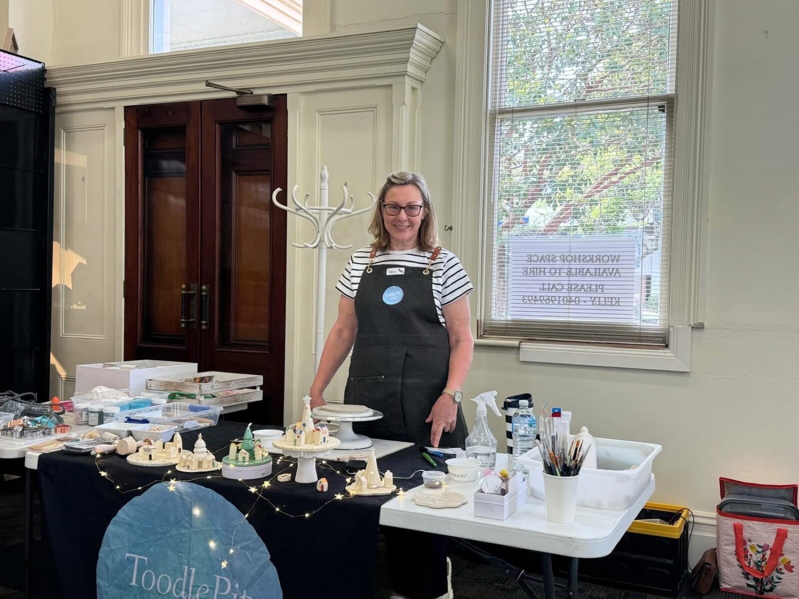 Dani Green at a workshop bench with hand-built ceramic village houses, glaze pots and brushes laid out for a class.